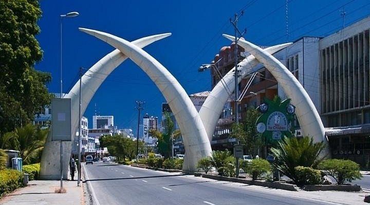 he Mombasa Tusks illuminated at night, with traffic passing underneath and palm trees lining Moi Avenue.
